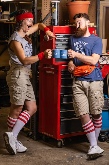 Gallery 1 - Two dads in khakis and t-shirts clink coffee mugs in the garage standing in front of a tool cart.